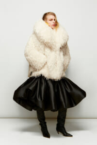 Model wearing an oversized cream shaggy faux-fur coat over a voluminous black skirt in a studio setting.