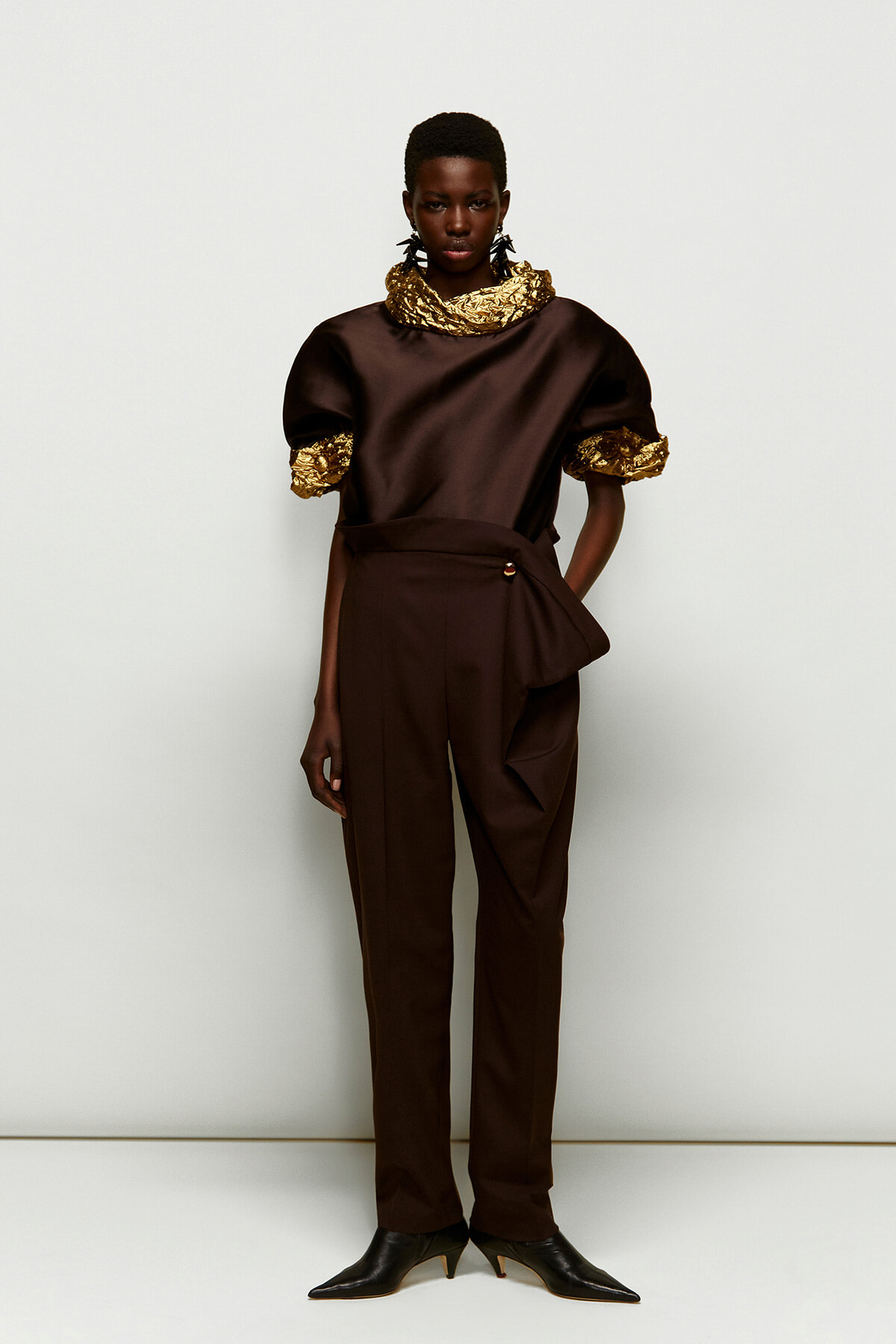 Model in a dark brown satin top with gold crinkled scarf and cuffs, paired with matching high-waisted pants and black heeled boots, fashion shoot