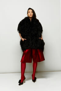 Full-length portrait of a person in a voluminous black feathered coat, red satin shorts, red tights, and black heels, posed against a white backdrop.