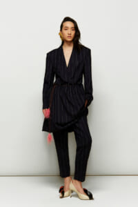Model wearing a navy pinstripe wrap blazer with matching tapered pants, belted at the waist with pink tassel accents, gold statement earring, standing against a pale backdrop.