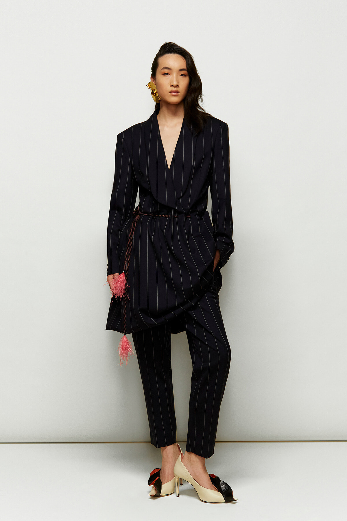 Model wearing a navy pinstripe wrap blazer with matching tapered pants, belted at the waist with pink tassel accents, gold statement earring, standing against a pale backdrop.