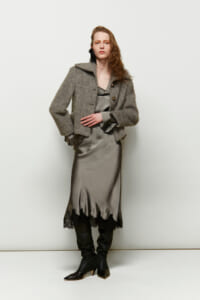Model wearing a gray textured coat over a satin gray slip dress with a lace hem, paired with black knee-high boots, standing against a light backdrop.