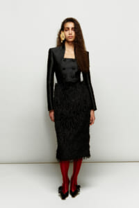 Model wearing a black, double-breasted blazer with a textured black midi skirt, red tights, and black bow-heeled shoes, plus a large gold earring.