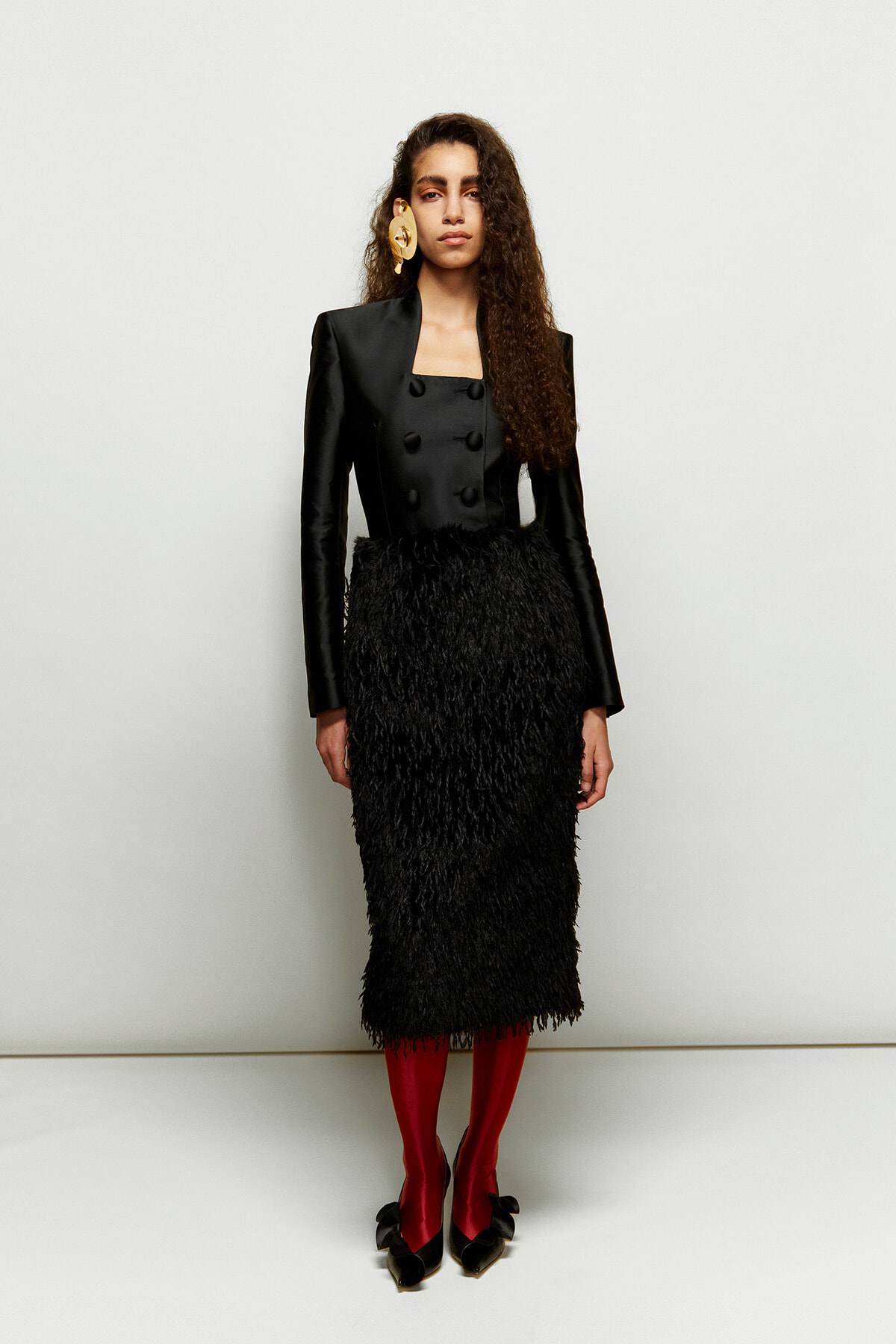 Model wearing a black, double-breasted blazer with a textured black midi skirt, red tights, and black bow-heeled shoes, plus a large gold earring.