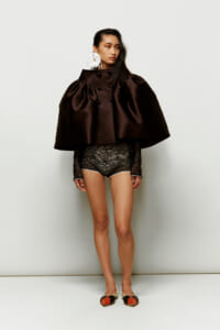 Model in a dark brown oversized cape with puffed sleeves, black lace shorts, and a large white feather earring, standing against a white backdrop.