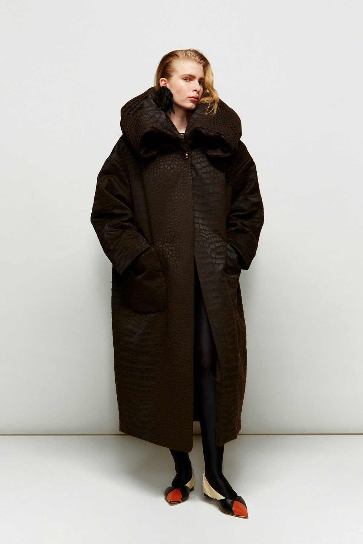 Model wearing a long dark brown textured coat with an oversized collar, hands in pockets, standing against a neutral backdrop.