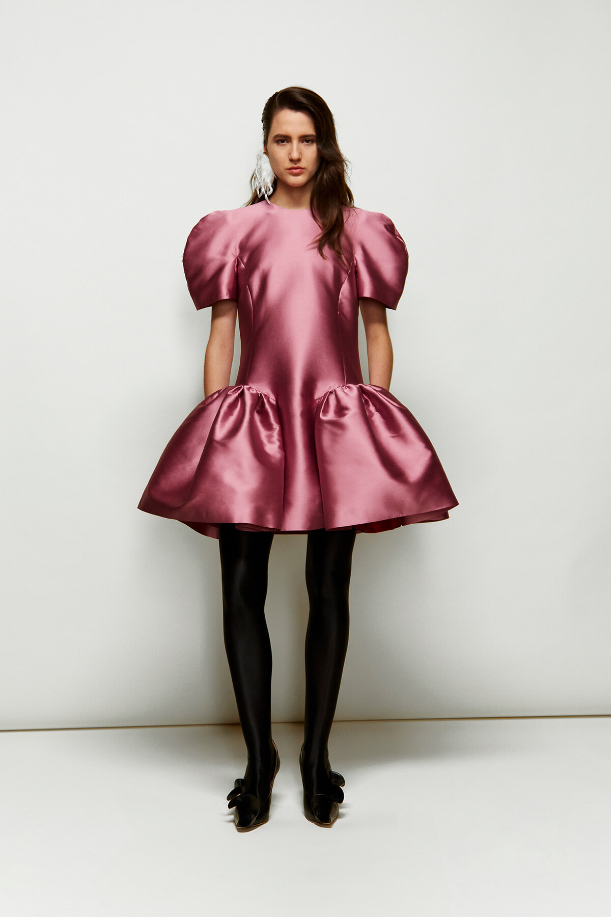 Model wearing a shiny pink satin dress with large puff sleeves and a voluminous skirt, standing against a light backdrop.