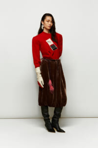 Fashion model wearing a red knit sweater with a patch, a voluminous brown velvet skirt, cream gloves, and tall black boots, posing against a light backdrop.