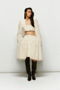 Model wearing a cream cropped top and high-waisted skirt with a fluffy cream coat and black knee-high boots, standing against a pale backdrop.