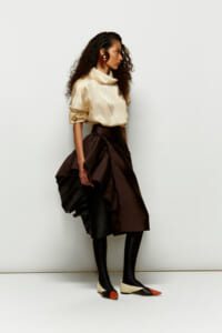 Model wearing a cream satin blouse and a layered brown asymmetric skirt with black tights and color-block flats, against a white backdrop with bold gold earrings.