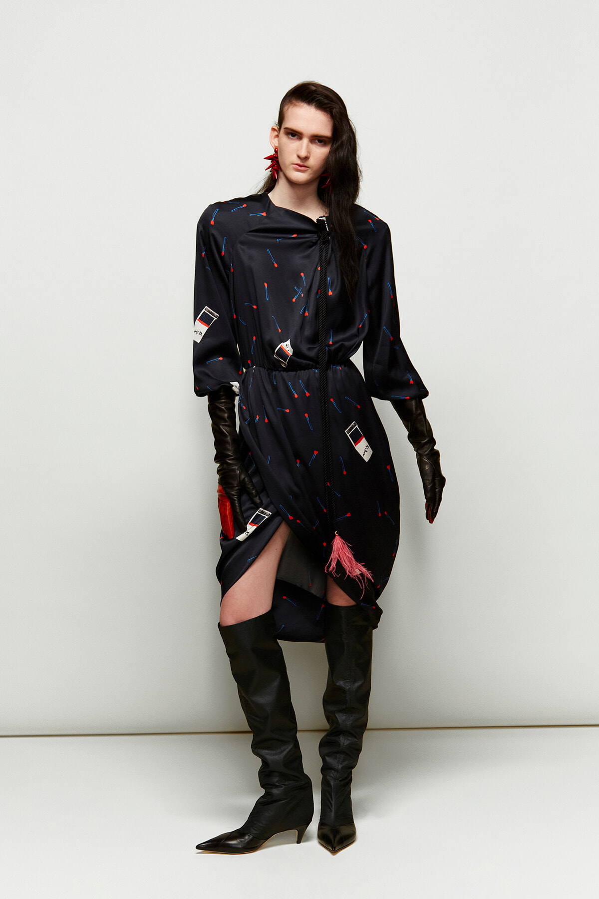 Model wearing a navy printed dress with a waist tie, pink tassel, red earrings, long brown gloves, and black knee-high boots.