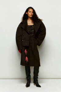 Woman in a dark textured overcoat with oversized silhouette, black outfit, thigh-high boots, and pink tassels hanging from the coat.