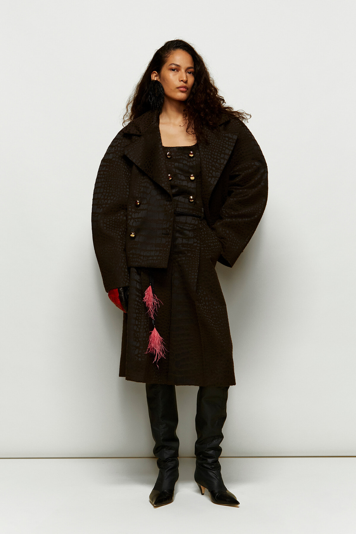 Woman in a dark textured overcoat with oversized silhouette, black outfit, thigh-high boots, and pink tassels hanging from the coat.