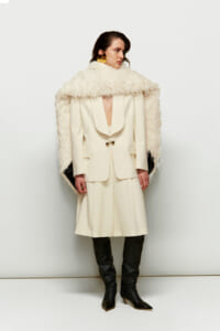 Model in an ivory tailored blazer and cape with a textured, shaggy white cape draped over shoulders, paired with black knee-high boots and gold hoop earrings.