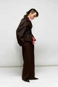 Side profile of a person wearing a dark textured oversized jacket, brown wide-leg pants, pink scarf, and large gold hoop earrings with a pink tassel belt.