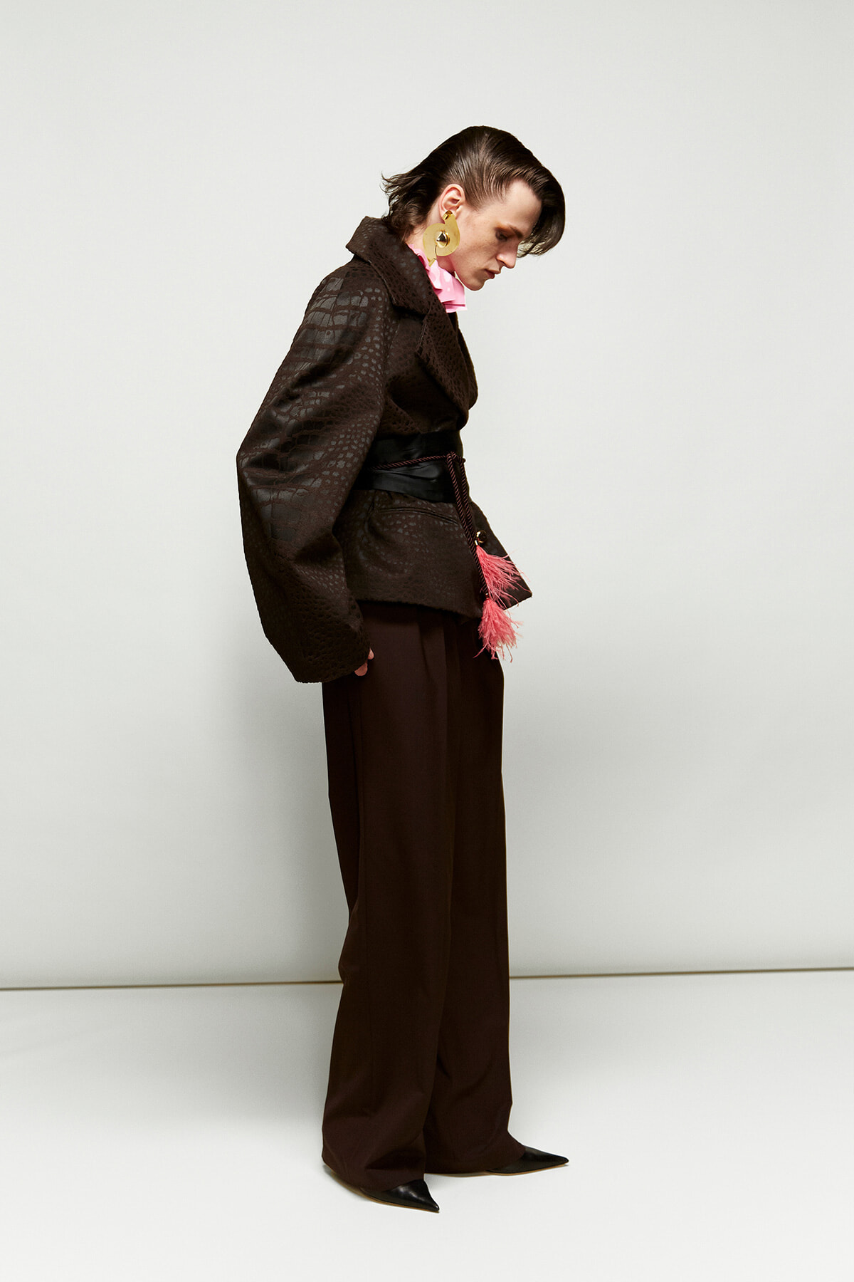 Side profile of a person wearing a dark textured oversized jacket, brown wide-leg pants, pink scarf, and large gold hoop earrings with a pink tassel belt.