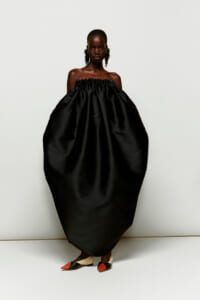 Model wearing an oversized off-shoulder black gown with smocked top, standing against a light background.