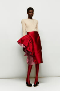 Model wearing a cream cardigan with a dramatic red satin asymmetric skirt that has pink lining and matching red tights, black bow heels.