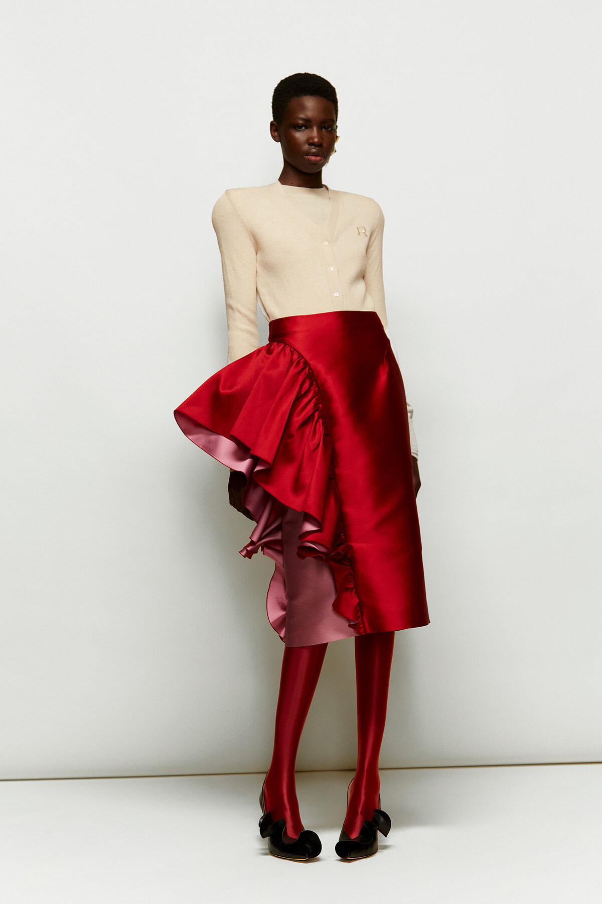 Model wearing a cream cardigan with a dramatic red satin asymmetric skirt that has pink lining and matching red tights, black bow heels.