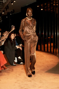 Model walking a runway in a bronze sequined suit with a deep V neckline, statement jewelry, and hands in pockets.