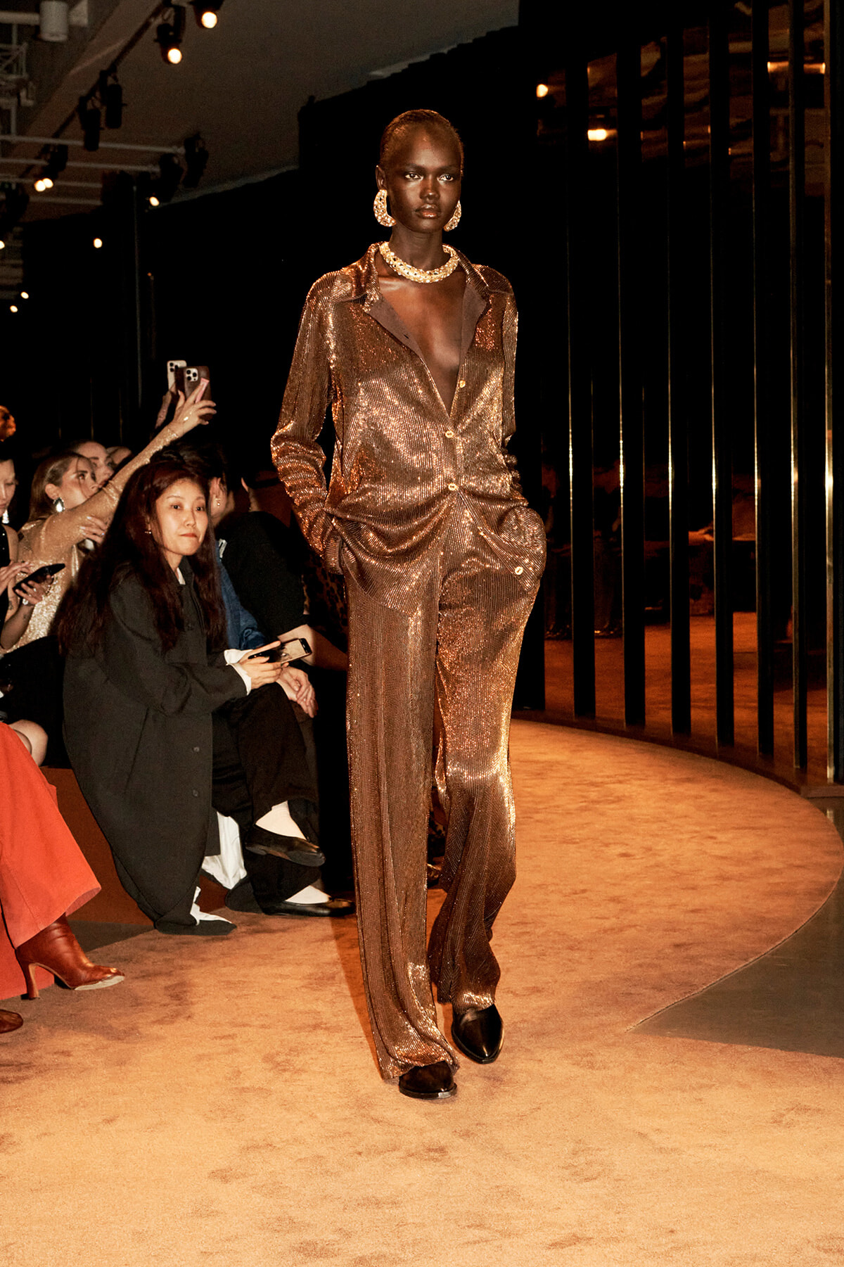 Model walking a runway in a bronze sequined suit with a deep V neckline, statement jewelry, and hands in pockets.