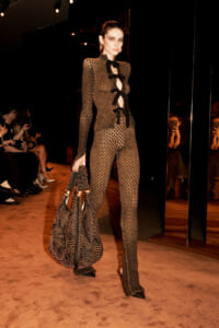 Model walks the runway in a sheer lattice jumpsuit with black bows, carrying a patterned large handbag as spectators watch nearby.