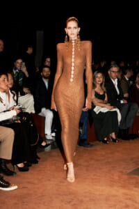 Model walking a runway in a bronze mesh, full-length dress with circular cutouts along the front, paired with statement earrings and nude heels; seated audience in the background.