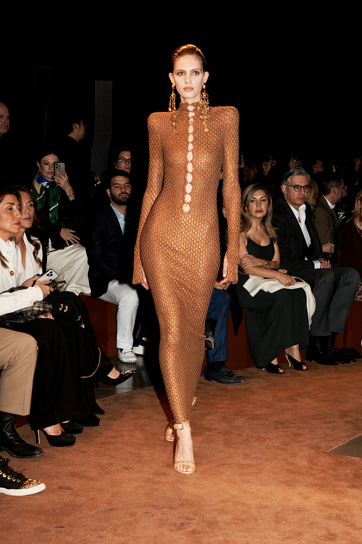Model walking a runway in a bronze mesh, full-length dress with circular cutouts along the front, paired with statement earrings and nude heels; seated audience in the background.