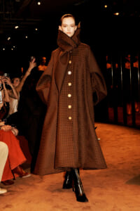Model on a runway wearing a voluminous brown plaid cape coat with a high oversized collar and gold buttons.