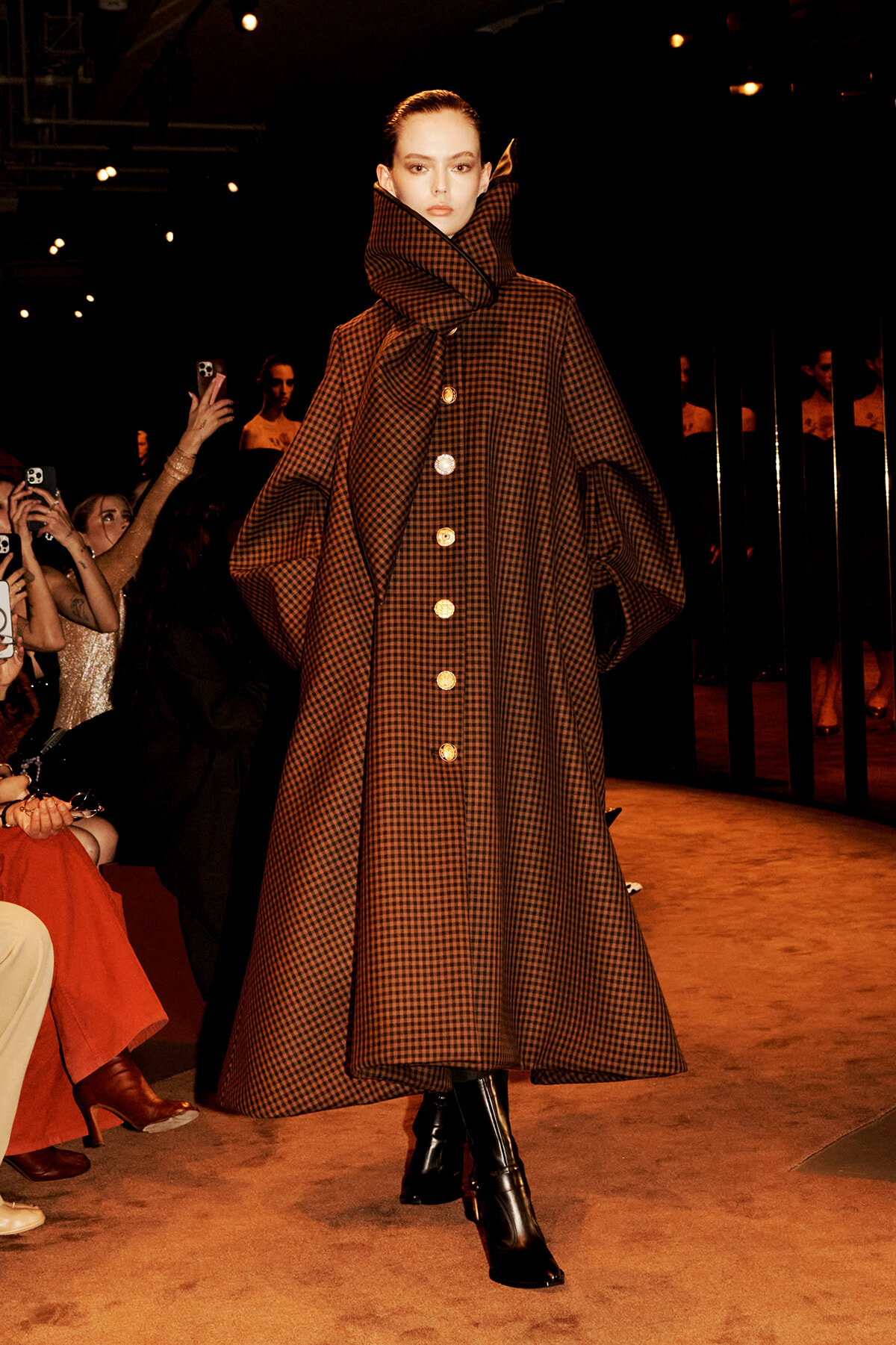 Model on a runway wearing a voluminous brown plaid cape coat with a high oversized collar and gold buttons.