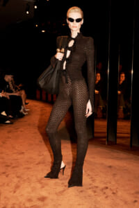 Model wearing a black textured jumpsuit with a lace-like pattern, holding a large black bag and wearing cat-eye sunglasses on a runway.