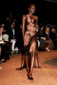 Model walks the runway in a metallic bronze, sculpted one-shoulder gown with diagonal pleats; audience watches from the sides in a dim venue.