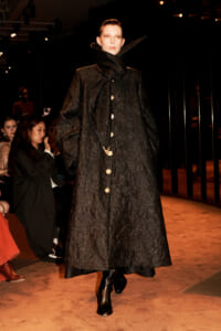 Model on a runway wearing a long black quilted coat with a high, oversized collar and gold buttons.