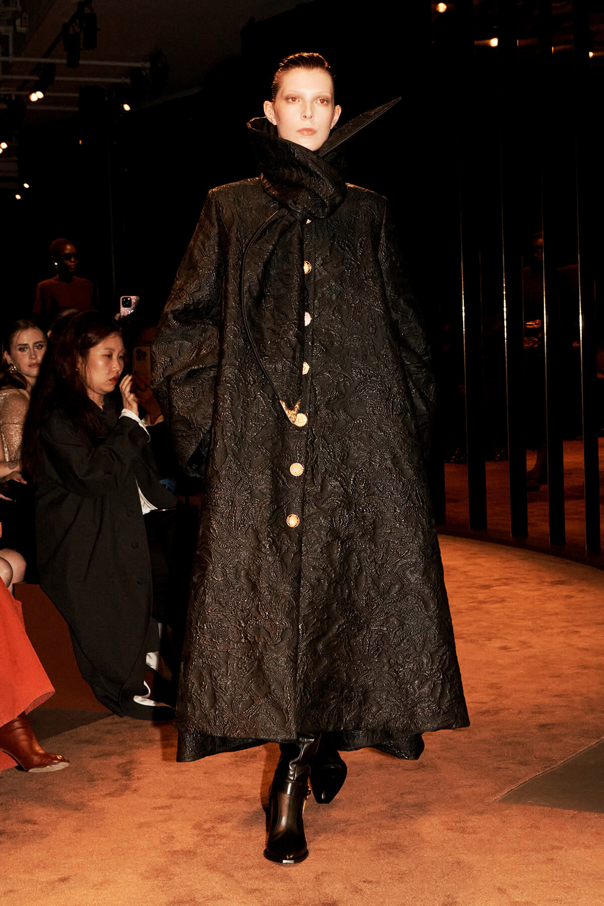 Model on a runway wearing a long black quilted coat with a high, oversized collar and gold buttons.