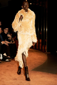 Model walking a fashion runway in a cream shaggy coat with a fringed dress and brown ankle boots; audience sits along the catwalk.