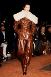 Model walks a fashion runway in a brown embossed suit with a large white fur collar, audience in the background.