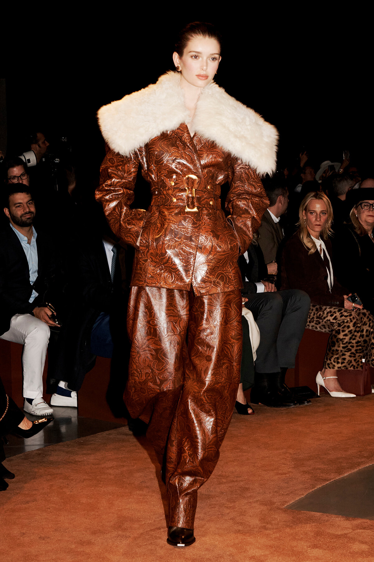 Model walks a fashion runway in a brown embossed suit with a large white fur collar, audience in the background.
