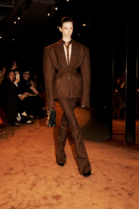 Model walks the runway in a brown checkered suit with oversized blazer and flared trousers, holding a black-gold handbag as audience watches.