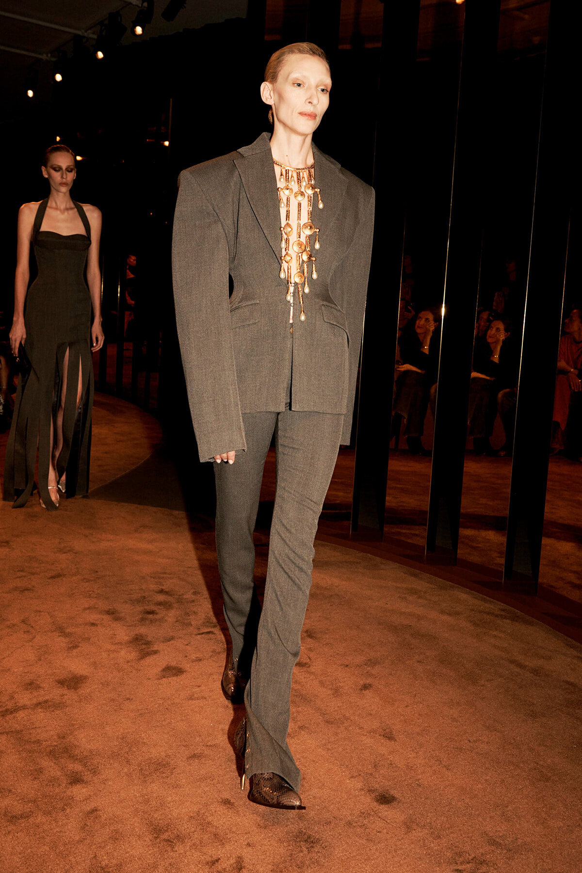 Model walking a fashion show in a tailored gray suit with a long, bold gold necklace; audience in the background.
