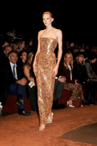 Model in a gold sequin strapless gown walking the runway at a fashion show, audience watching.