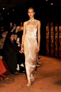 Model walking the runway in a sequined peach halter gown, audience photographing in the background.