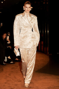 Model on a runway wearing a cream textured pantsuit with oversized shoulders, pearl earrings, and a jeweled clutch.