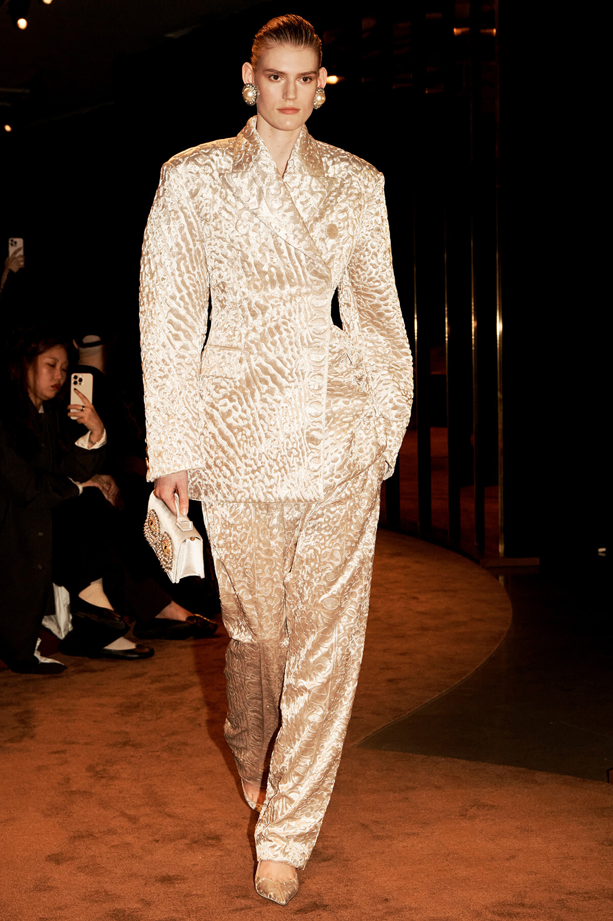 Model on a runway wearing a cream textured pantsuit with oversized shoulders, pearl earrings, and a jeweled clutch.