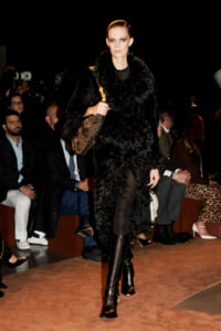 Model walks runway in a black fur coat, thigh-high boots, and a studded handbag at a fashion show.