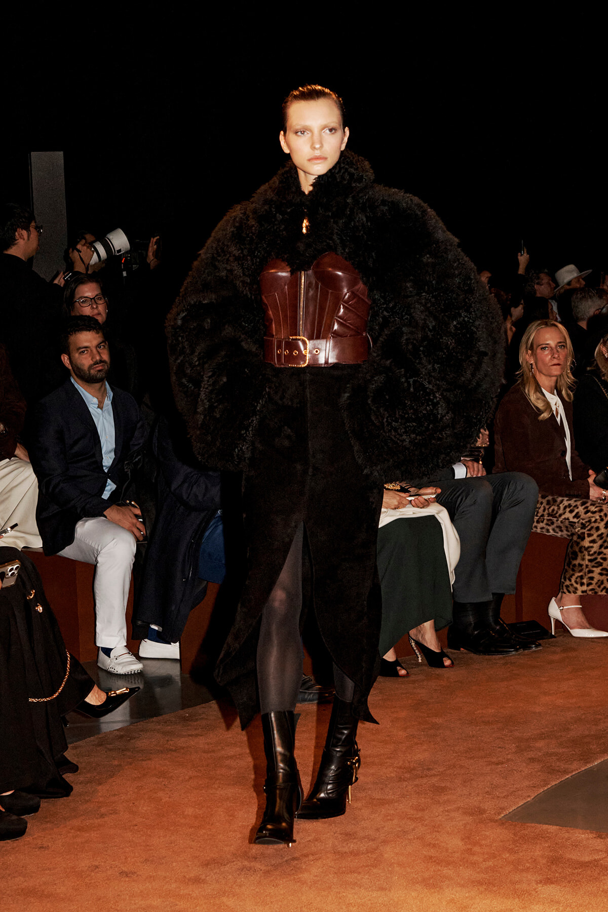 Model walking the runway in a voluminous black fur coat belted with a wide burgundy leather corset and black knee-high boots, audience in background.