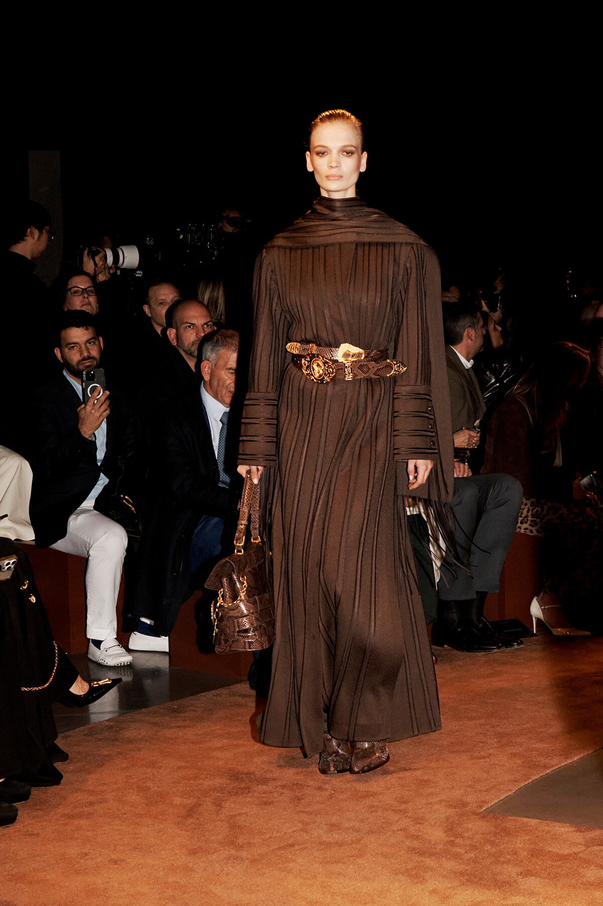 Model in a brown pleated gown with a wide gold belt walking a runway; audience seated along the sides.