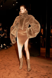 Model walking a fashion show in a oversized brown fur coat, a high-slit bronze skirt, tan suede boots, and a beige handbag, with audience in the background.