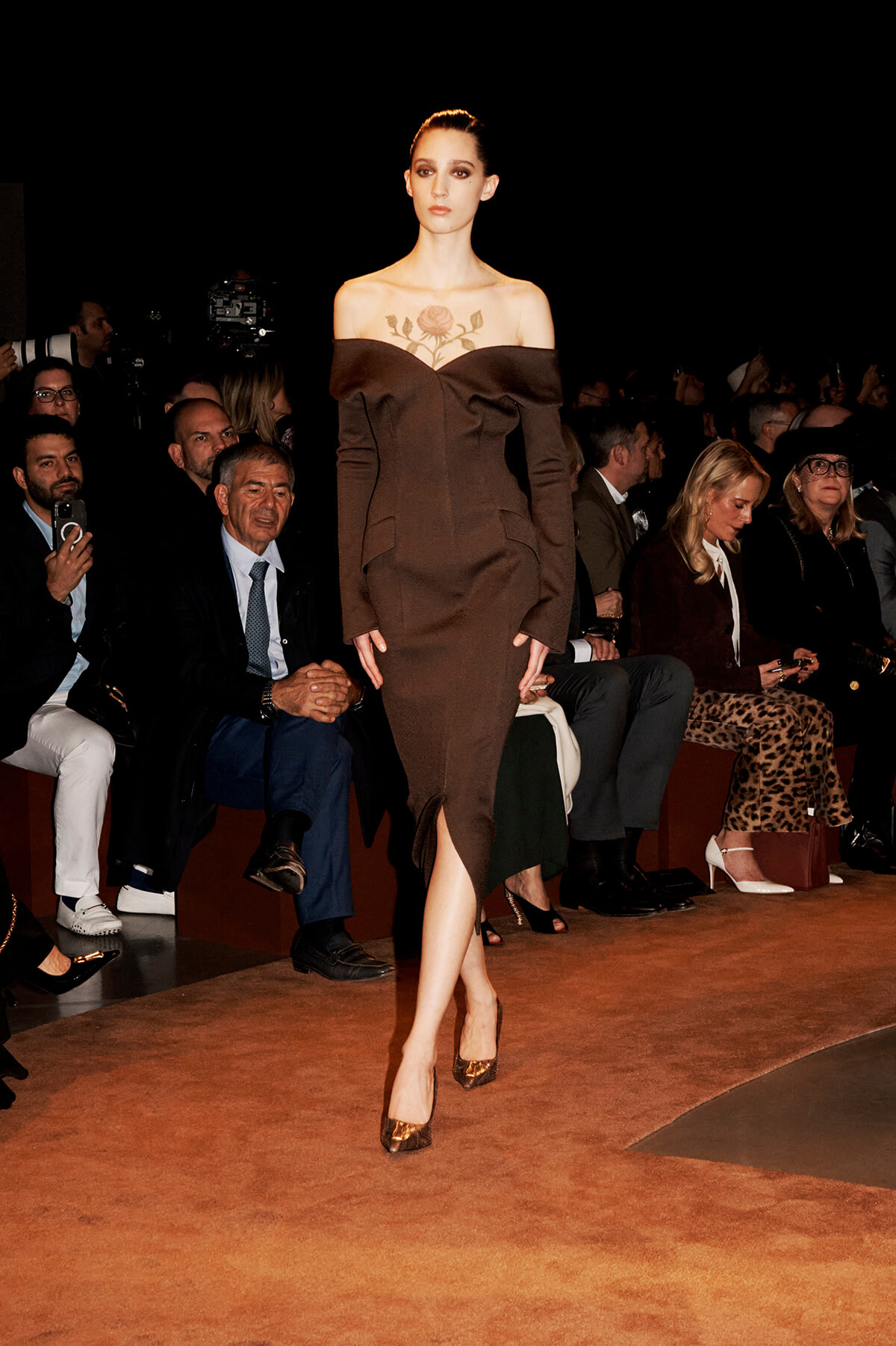 Model walking a fashion show in a brown off-shoulder dress; visible rose tattoo on chest, audience seated along runway behind her.