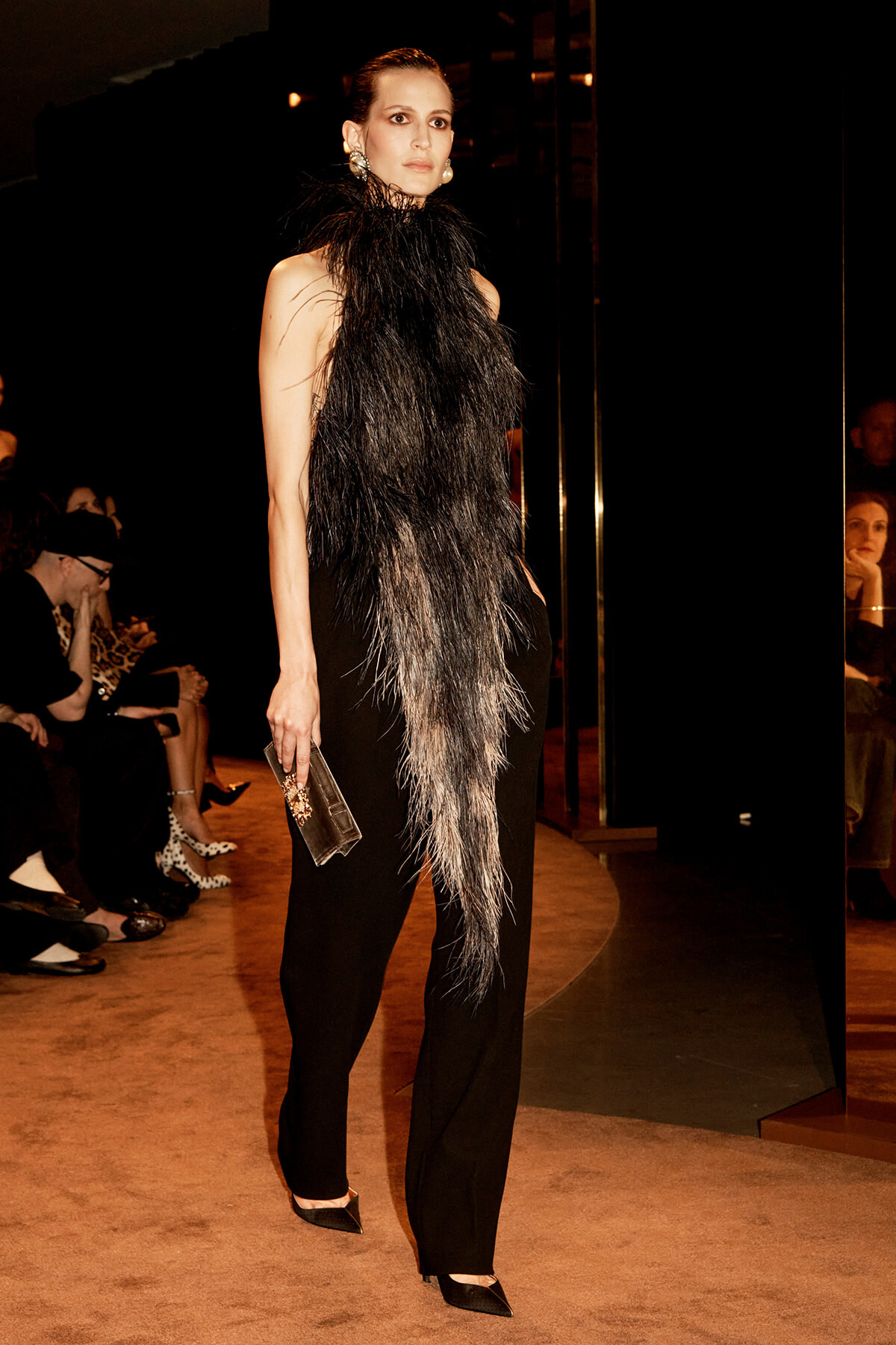 Model on a runway wearing a black feathered high-neck top and black pants, holding a rectangular clutch.