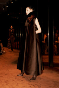 Model on a runway wearing a long black satin dress with a fur scarf and snakeskin boots, audience in dim lighting behind a reflective wall.